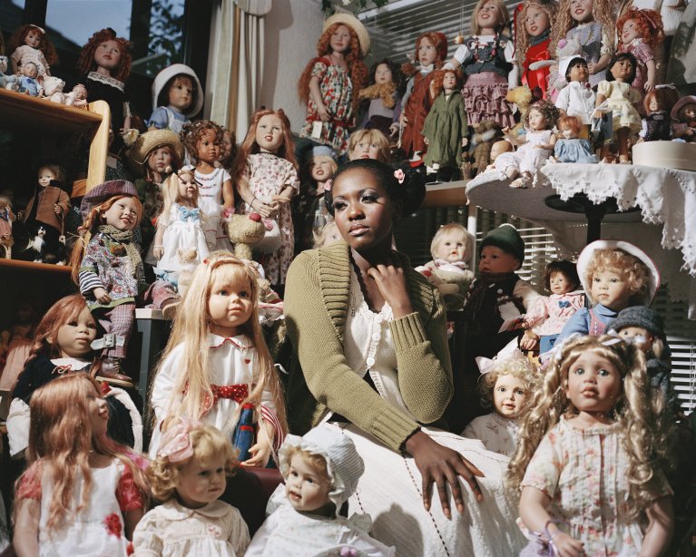 Artwork by Maziar Moradi, Black Woman sitting, surounded by white dolls