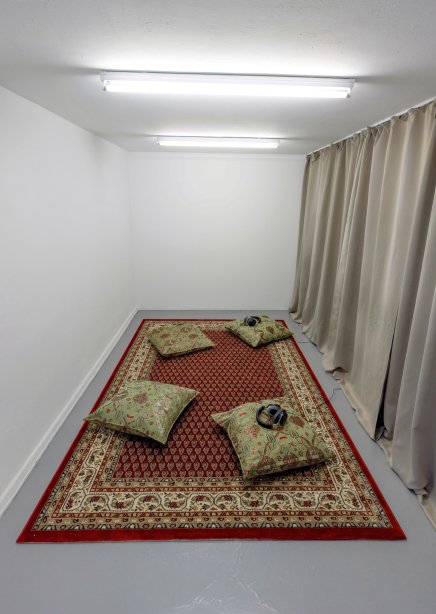 small room with oriental carpet, curtains and four cushions and a sound installation with headphones